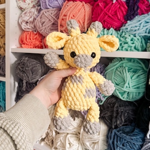 May include: A crocheted giraffe toy in yellow and grey. The giraffe has a textured, plush appearance with grey spots, ears, and feet. The background shows shelves filled with various colours of yarn.