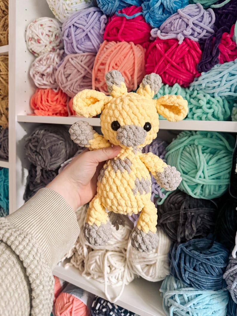 May include: A handmade yellow and gray crochet giraffe toy. The giraffe has a textured, plush appearance with gray spots, ears, and feet. The background features shelves filled with various colors of yarn.
