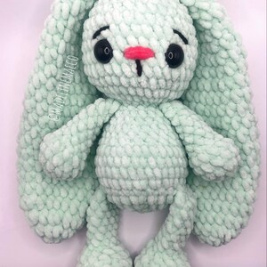 May include: A soft, crocheted green bunny with long ears and a pink nose. The bunny has black eyes and is sitting on a white background.