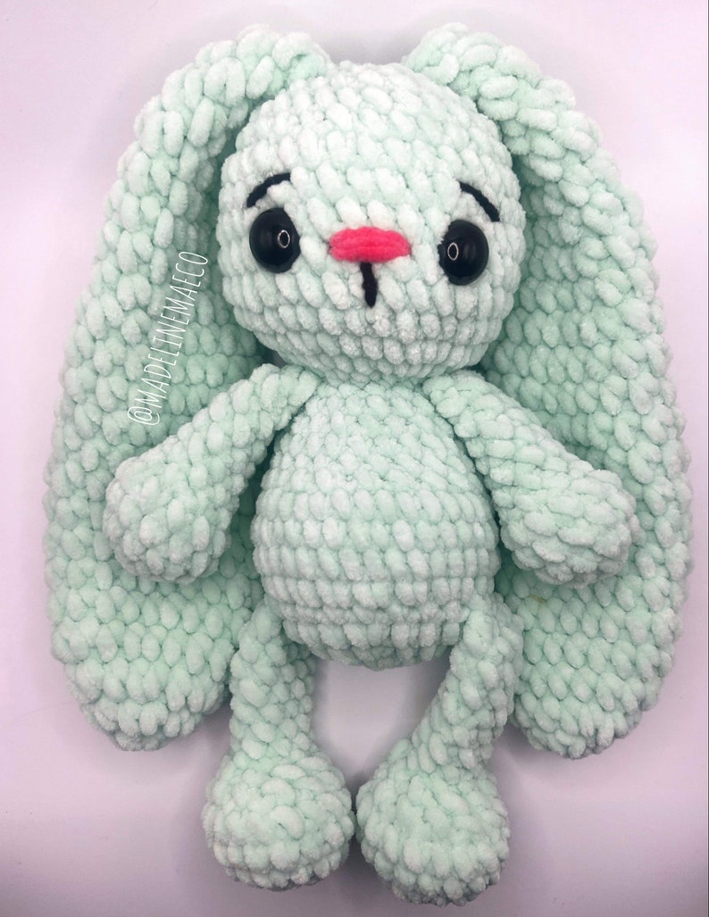 May include: A soft, crocheted green bunny with long ears and a pink nose. The bunny has black eyes and is sitting on a white background.