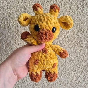 May include: A crocheted giraffe stuffed animal with brown spots and a brown nose. The giraffe is yellow and has black eyes.