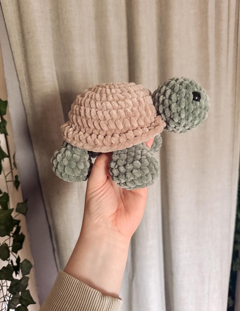 May include: A handmade crochet turtle in soft, plush yarn. The turtle features a light brown shell, a green head, and green feet. The turtle is a cuddly, whimsical toy.
