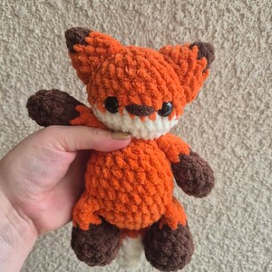 May include: A handmade orange and brown crochet fox toy with black eyes and a white muzzle. The fox has a fluffy texture and is being held up by a hand. The fox's ears and paws are brown, and the body is orange.