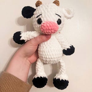 May include: A white crocheted cow plush toy with black spots, brown ears, and a pink nose. The toy has black eyes and is standing on its four legs.