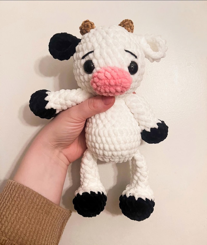 May include: A white crocheted cow plush toy with black spots, brown ears, and a pink nose. The toy has black eyes and is standing on its four legs.