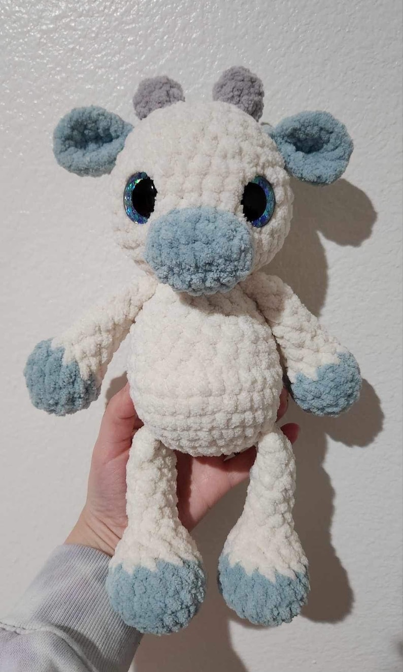 May include: A crocheted stuffed animal in the shape of a giraffe with white fur, blue ears, and blue hooves. The giraffe has large blue eyes with glitter.
