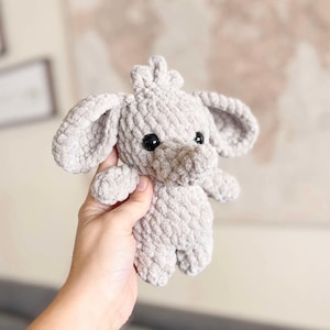 Könnte beinhalten: Ein handgehäkelter, hellgrauer Elefant mit schwarzen Sicherheitsaugen und großen, schlaffen Ohren. Das Plüschtier wird in einer Hand gehalten und zeigt sein weiches, strukturiertes Aussehen. Der Elefant hat einen kleinen Rüssel und ein kronenartiges Detail auf dem Kopf.