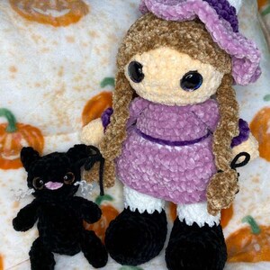 May include: A handmade crochet doll with brown hair, a purple dress, and a matching hat. The doll has large, expressive eyes and is accompanied by a small black crochet cat. The background features a pumpkin-themed pattern.