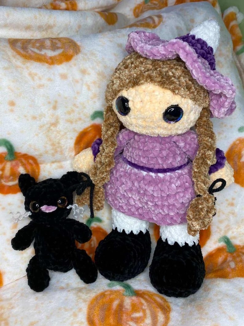May include: A handmade crochet doll with brown hair, a purple dress, and a matching hat. The doll has large, expressive eyes and is accompanied by a small black crochet cat. The background features a pumpkin-themed pattern.
