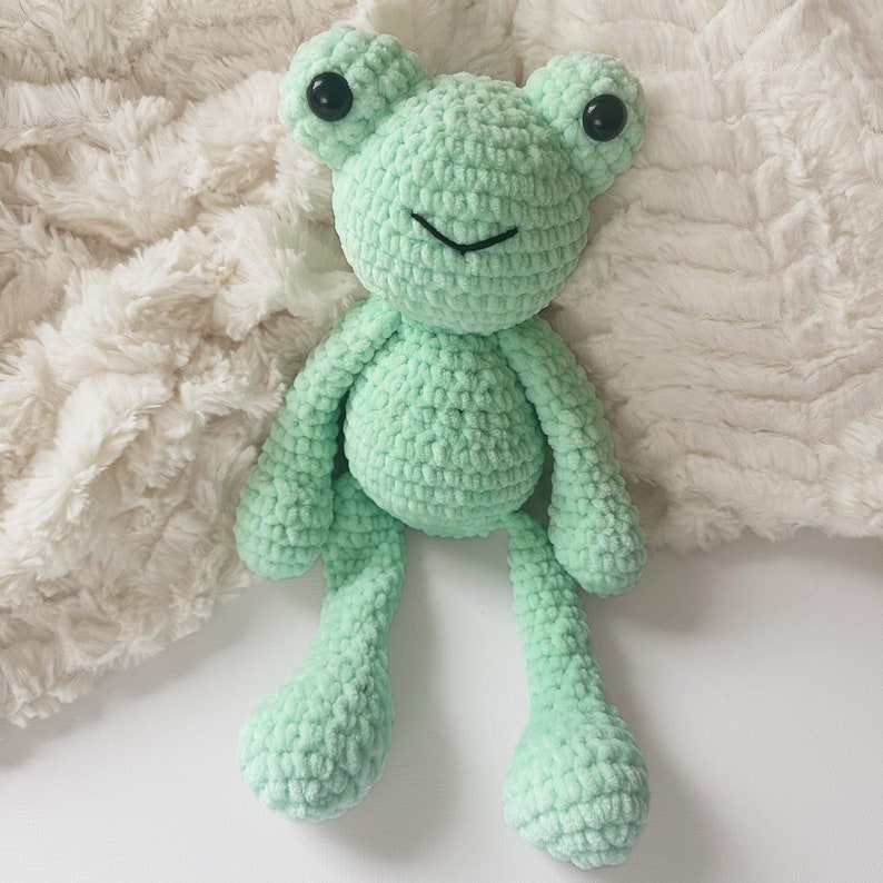 May include: A green crocheted frog toy with black eyes. The frog is sitting on a white fluffy surface.