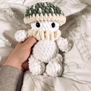 Pode incluir: Um brinquedo de pelúcia branco feito à mão em forma de cogumelo, com uma touca verde e branca, brânquias creme e olhos pretos. O brinquedo macio e fofinho é segurado em uma mão e repousa sobre uma colcha com estampa floral.