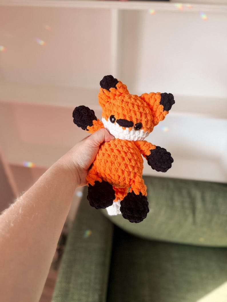 May include: A handmade orange and black crochet fox toy with white detailing. The fox has black eyes and a black nose. The fox is a stuffed animal made from soft, textured yarn. A cute and cuddly toy.