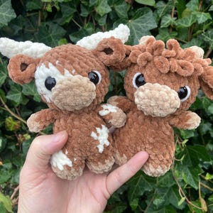 May include: Two crocheted plush toys shaped like brown and white Highland cows. The cows have curly fur and big eyes.