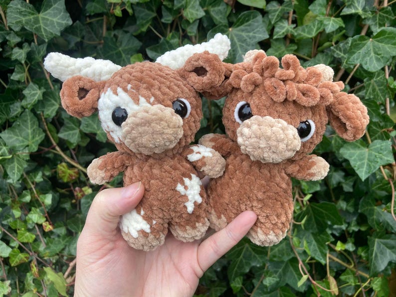 May include: Two crocheted plush toys shaped like brown and white Highland cows. The cows have curly fur and big eyes.