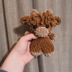 May include: A brown crocheted stuffed animal resembling a moose with large, brown eyes. The animal is held in a person's hand.