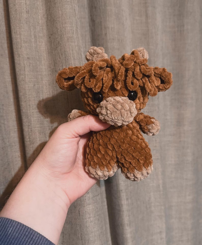May include: A brown crocheted stuffed animal resembling a moose with large, brown eyes. The animal is held in a person's hand.