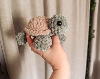 Puede incluir: Una tortuga de ganchillo hecha a mano en tonos verde salvia y rosa empolvado. La tortuga tiene un caparazón y un cuerpo texturizados, con un ojo negro. Este juguete de peluche suave es adecuado para una habitación infantil o como objeto decorativo.