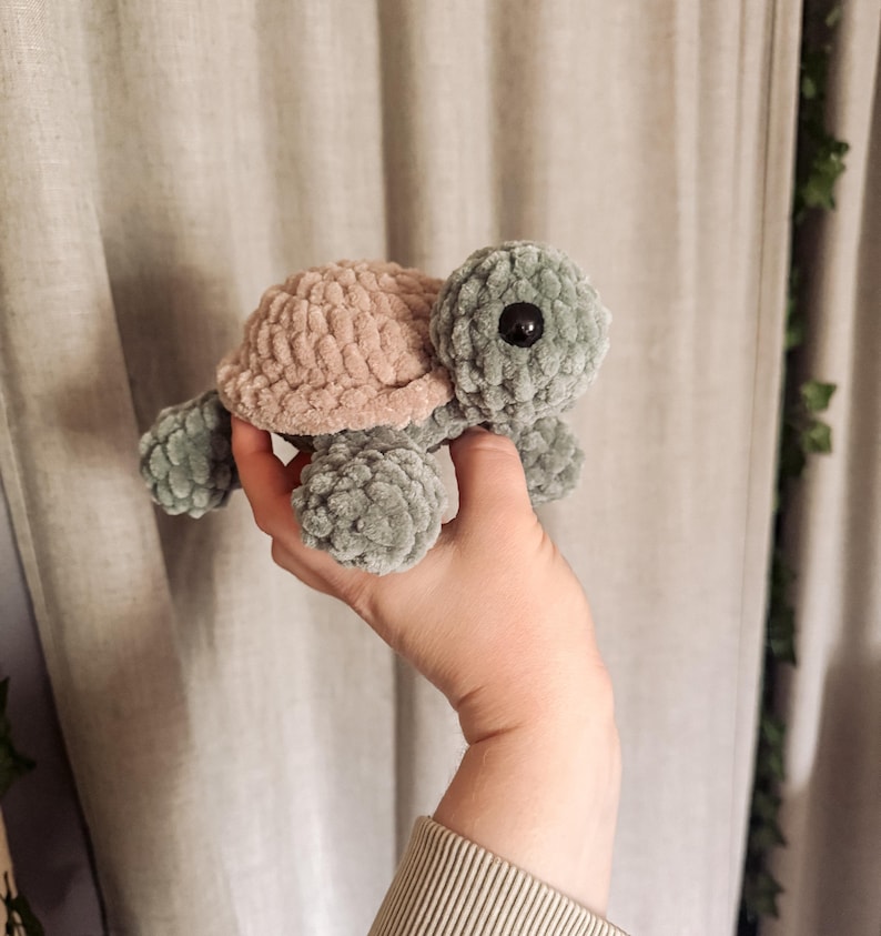 May include: A handmade crochet turtle in shades of sage green and dusty rose. The turtle has a textured shell and body, with a black eye. The plush toy is soft and cuddly, perfect for a child's room or as a decorative item.