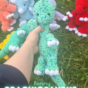 May include: A plush crochet Brachiosaurus toy in variegated shades of green and blue with white claws. The text Benny BRACHIOSAURUS is printed at the bottom. Other colourful plush dinosaur toys are visible in the background.
