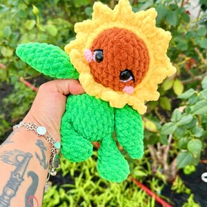 K&ouml;nnte beinhalten: Ein geh&auml;keltes Pl&uuml;sch-Sonnenblumenspielzeug mit gr&uuml;nem K&ouml;rper und gelben Bl&uuml;tenbl&auml;ttern. Die Sonnenblume hat ein braunes Gesicht mit schwarzen Augen und rosa Wangen.