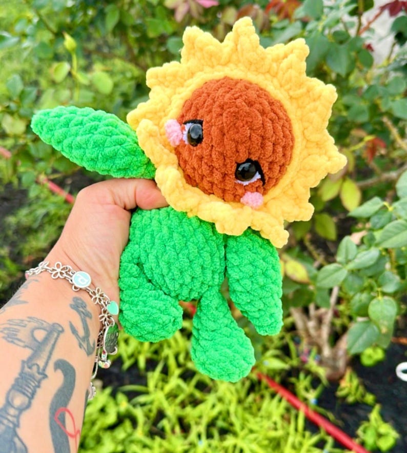 K&ouml;nnte beinhalten: Ein geh&auml;keltes Pl&uuml;sch-Sonnenblumenspielzeug mit gr&uuml;nem K&ouml;rper und gelben Bl&uuml;tenbl&auml;ttern. Die Sonnenblume hat ein braunes Gesicht mit schwarzen Augen und rosa Wangen.