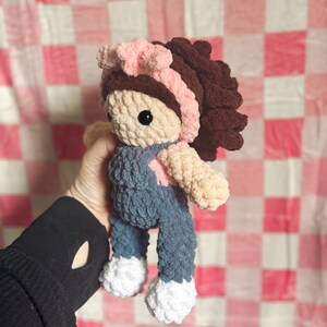 May include: A crocheted doll with brown hair, a pink headband, and blue overalls. The doll is holding a pink heart in its hand.