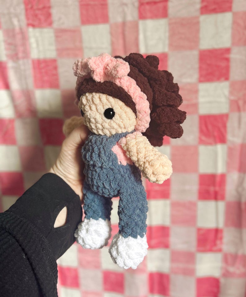 May include: A crocheted doll with brown hair, a pink headband, and blue overalls. The doll is holding a pink heart in its hand.