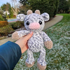 May include: A crocheted plush toy cow with a white and gray speckled body, brown ears, and a pink nose. The toy is being held in a hand against a snowy background.