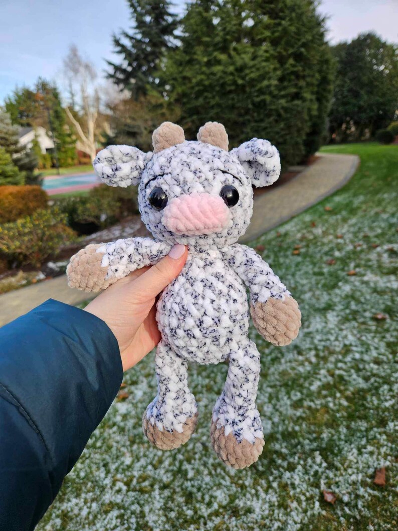 May include: A crocheted plush toy cow with a white and gray speckled body, brown ears, and a pink nose. The toy is being held in a hand against a snowy background.