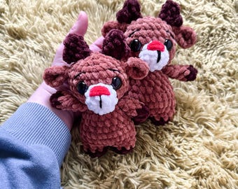 May include: Two brown crocheted reindeer toys with white faces and red noses. The reindeer are sitting on a fluffy beige surface.
