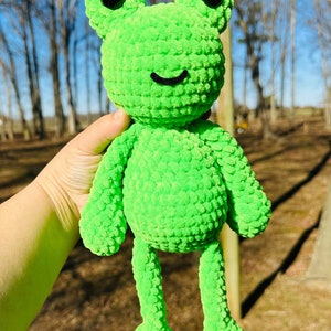May include: A bright green crocheted frog plush toy with black eyes and a smiling mouth. The frog is sitting in a person's hand.