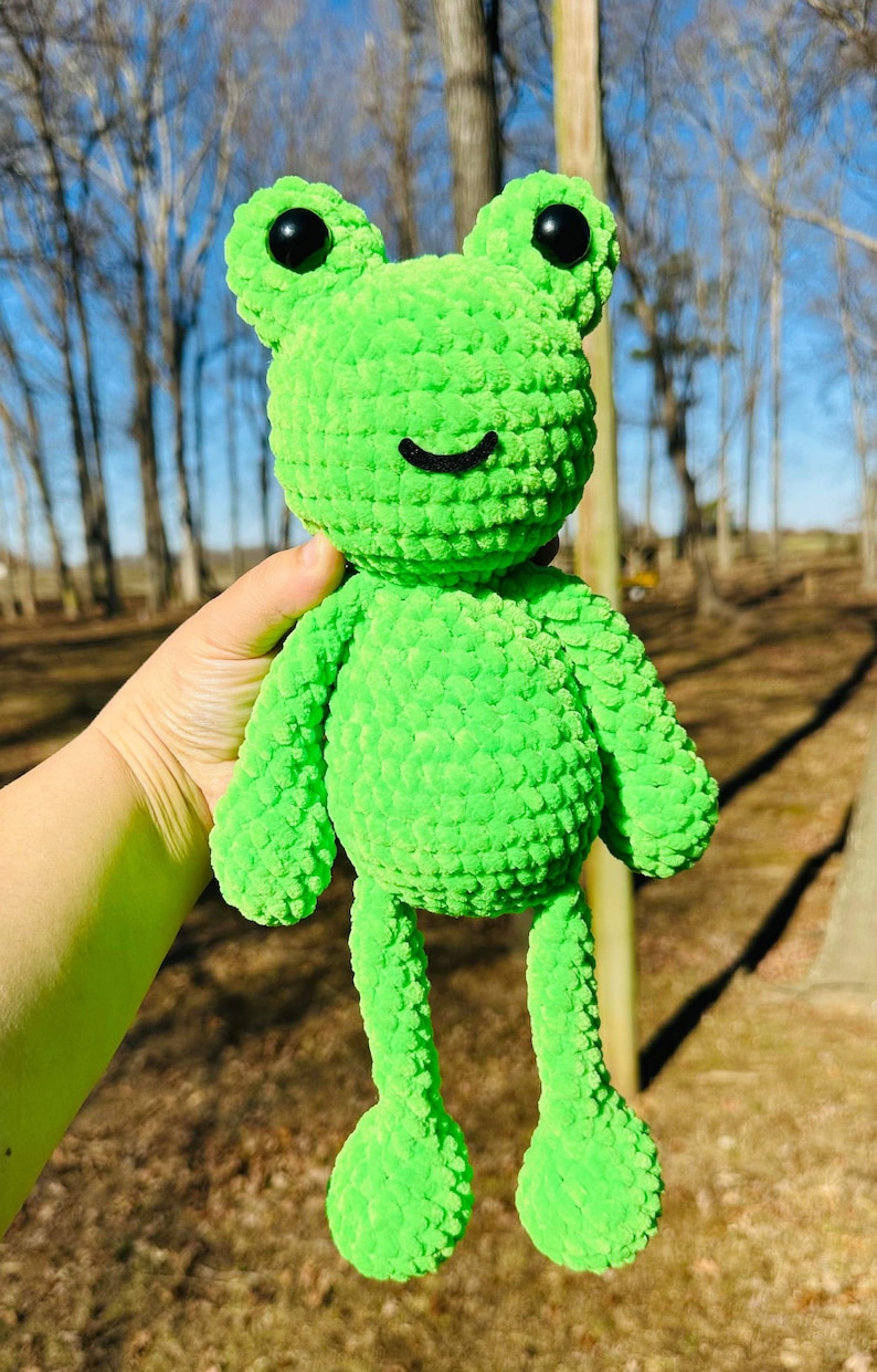 May include: A bright green crocheted frog plush toy with black eyes and a smiling mouth. The frog is sitting in a person's hand.
