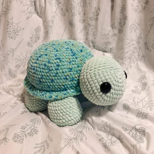Puede incluir: Un peluche de tortuga hecho a mano con ganchillo. La tortuga tiene un caparazón moteado en verde azulado y azul, una cabeza verde claro y cuatro patas a juego. Los ojos son grandes, negros y redondos. El peluche está sobre una superficie blanca con estampado floral.