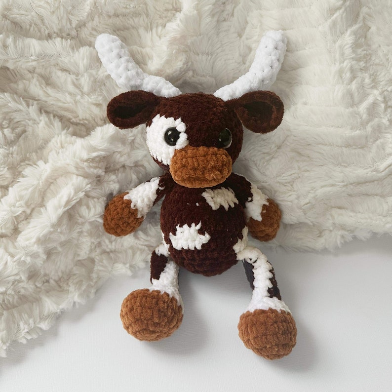 May include: A handmade crochet cow plushie with brown, white, and tan details. The stuffed animal has large black eyes, white horns, and is laying on a white, textured surface. The cow's body is a mix of brown and white.