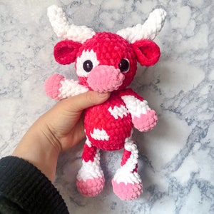 May include: A handmade plush cow toy in shades of red, white, and pink. The cow has large black eyes, white horns, and white patches on its body. The toy is made of soft, textured yarn, and is designed to be cuddly.