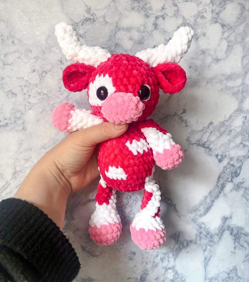 May include: A handmade plush cow toy in shades of red, white, and pink. The cow has large black eyes, white horns, and white patches on its body. The toy is made of soft, textured yarn, and is designed to be cuddly.
