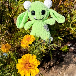 May include: A green crocheted plush toy with white ears and black eyes. The toy is sitting in a bed of yellow flowers.