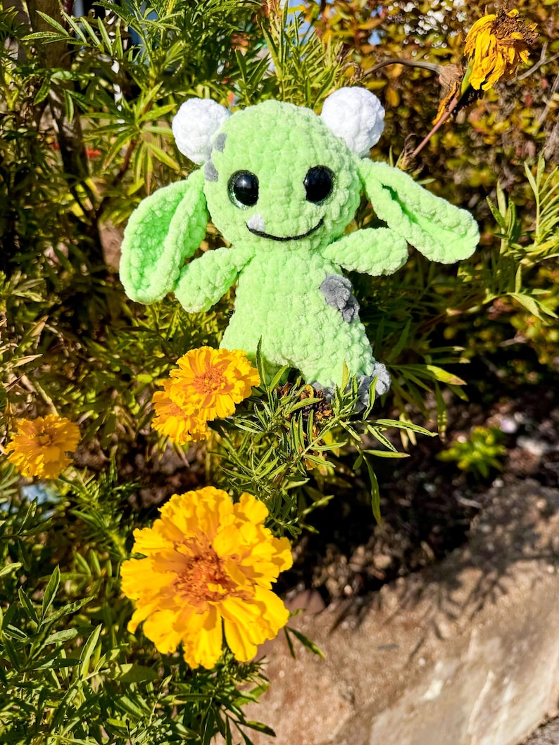 May include: A green crocheted plush toy with white ears and black eyes. The toy is sitting in a bed of yellow flowers.