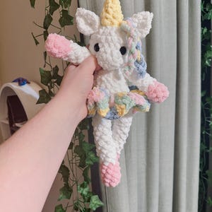 May include: A handmade white crochet unicorn doll with a yellow horn, pink hooves, and a colorful tutu. The unicorn has black eyes and is holding its arms out. The doll is made of soft, textured yarn.