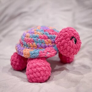 May include: A handmade crochet turtle in shades of pink, blue, orange, and purple. The turtle has a textured shell and body, with a pink head and legs. The eyes are black. This soft toy is perfect for a child's room.