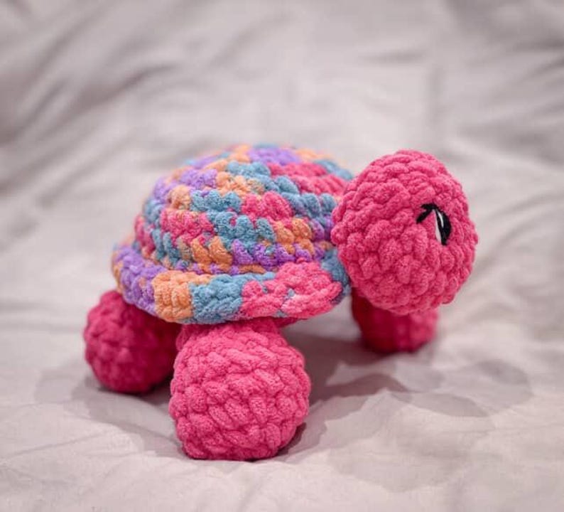 May include: A handmade crochet turtle in shades of pink, blue, orange, and purple. The turtle has a textured shell and body, with a pink head and legs. The eyes are black. This soft toy is perfect for a child's room.