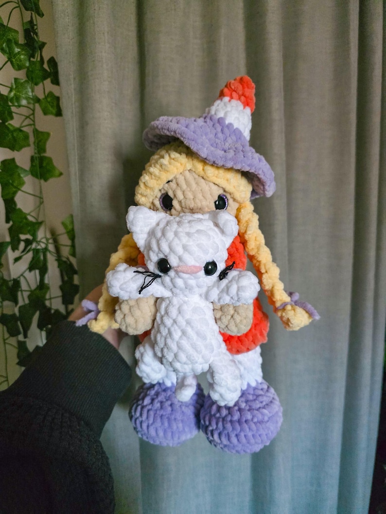 May include: A handmade crochet doll with blonde hair in braids, wearing a purple hat and holding a white cat. The doll has an orange and red outfit with purple shoes. The cat has black whiskers and pink accents.