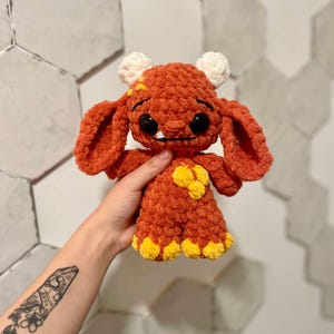 May include: A crocheted plush toy in the shape of a monster. The monster is orange with yellow accents and white eyes. It has a small mouth with two teeth and a slightly sad expression.