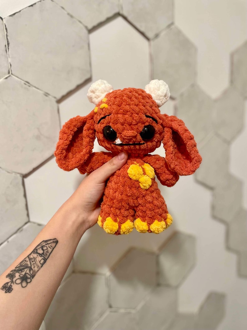 May include: A crocheted plush toy in the shape of a monster. The monster is orange with yellow accents and white eyes. It has a small mouth with two teeth and a slightly sad expression.