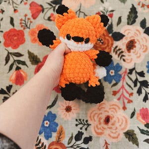 May include: A crocheted fox toy in vibrant orange, black, and white. The handmade plush animal features a rounded body, pointed ears, and a friendly expression. The fox is held against a floral patterned background.