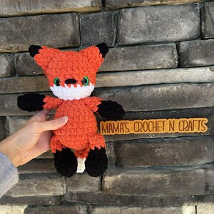 May include: A crocheted fox toy in orange with black tips on the ears and paws. It has a white collar and green eyes. The text "MAMA'S CROCHET N CRAFTS" is on a wooden sign.