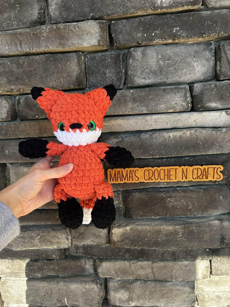 May include: A crocheted fox toy in orange with black tips on the ears and paws. It has a white collar and green eyes. The text "MAMA'S CROCHET N CRAFTS" is on a wooden sign.