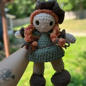 May include: A handmade crochet doll with a whimsical witch design. The doll features a green dress, brown hat, and orange hair. The doll's face is a light beige color, and it has large black eyes. The doll is approximately 8 inches tall.