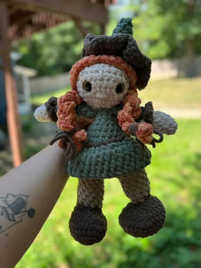 May include: A handmade crochet doll with a whimsical witch design. The doll features a green dress, brown hat, and orange hair. The doll's face is a light beige color, and it has large black eyes. The doll is approximately 8 inches tall.