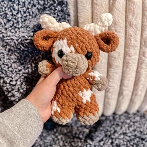 May include: A brown and white crocheted stuffed animal cow with a white star pattern. The cow has a brown nose and black eyes.